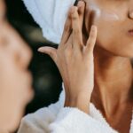 crop ethnic woman applying cream on face against mirror