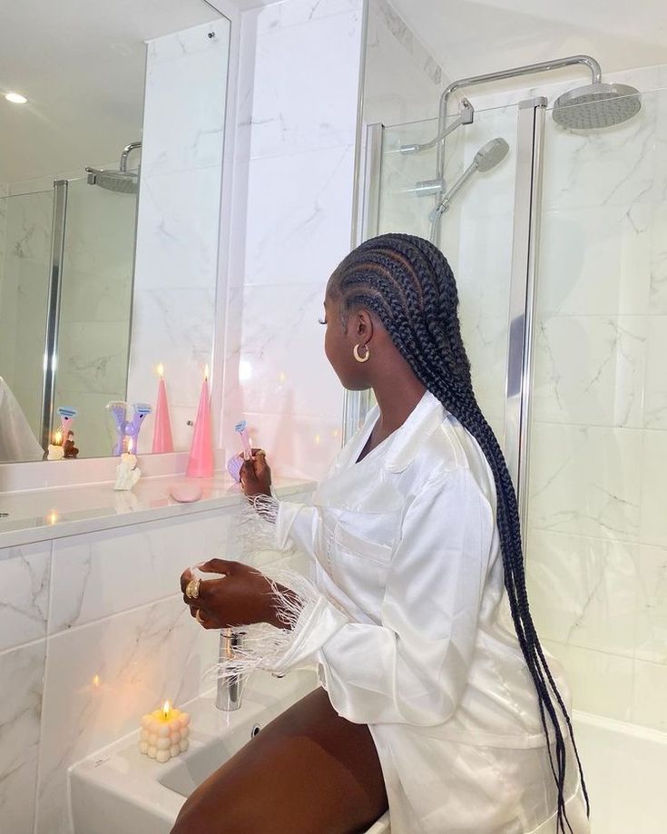 A woman sitting on the edge of a bathtub in a luxurious bathroom, surrounded by candles and soft lighting, wearing a white silk robe with feathered sleeves, creating a serene and indulgent self-care ambiance.