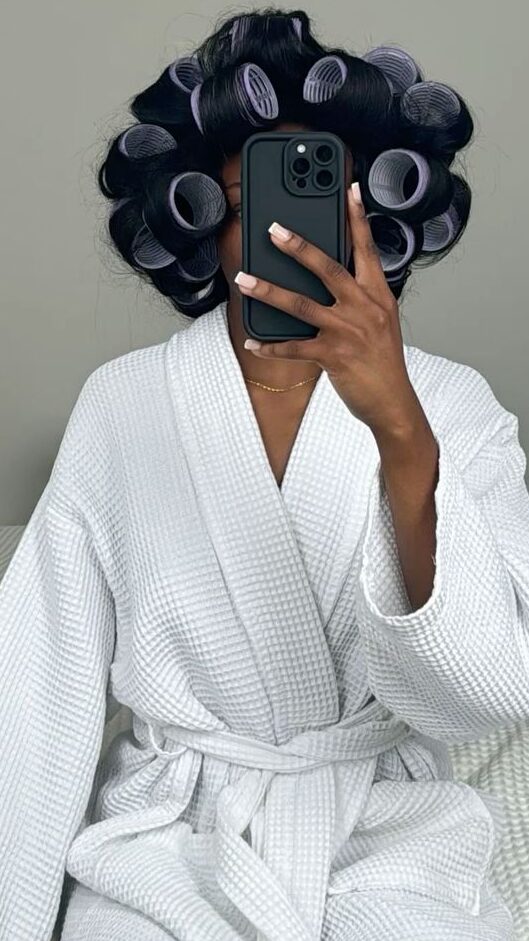 Person wearing a cozy white waffle robe with hair rollers, holding a phone to mirror. The setting exudes a relaxing self-care vibe with a touch of understated luxury.