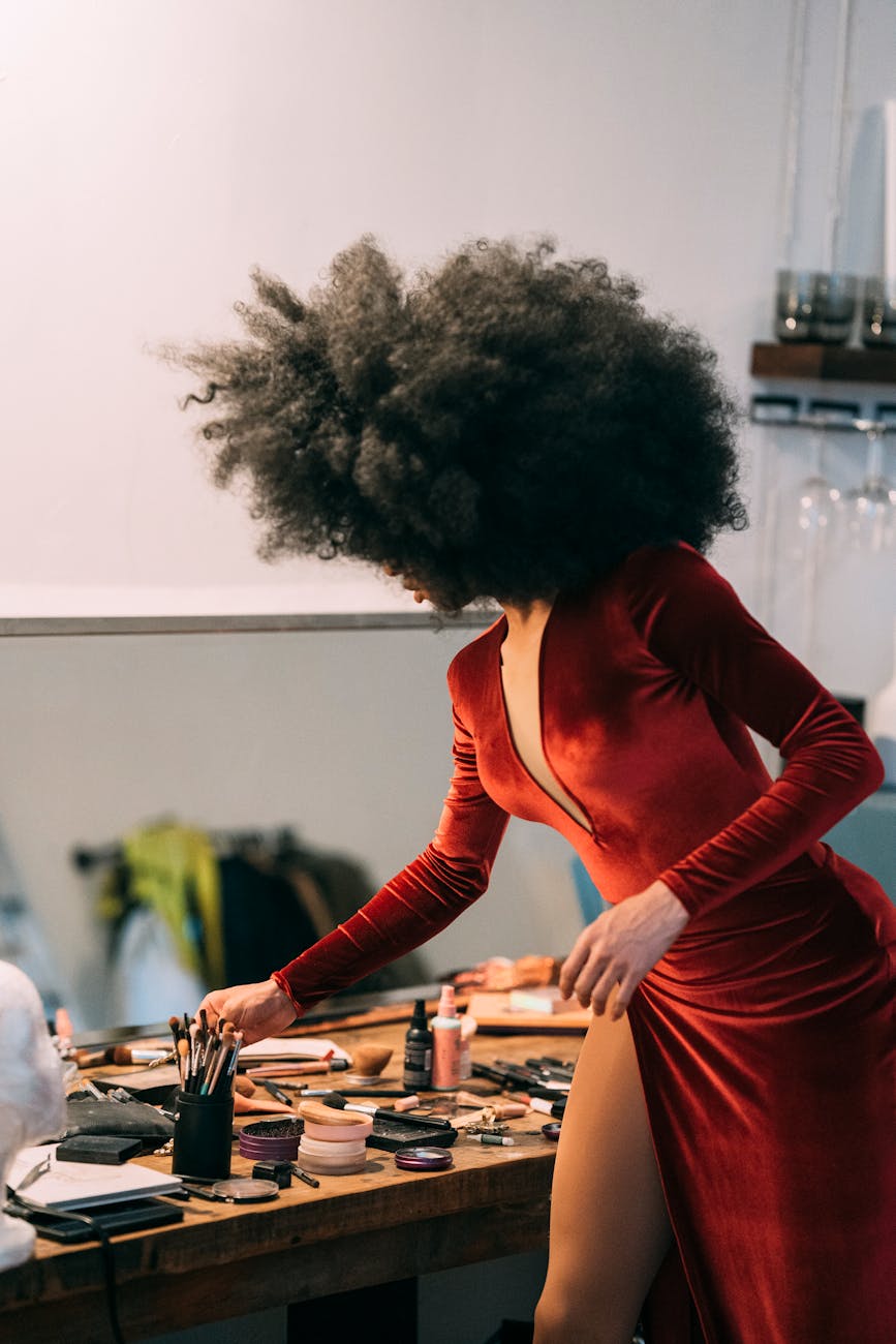 black model in stylish dress standing near table with cosmetics in room